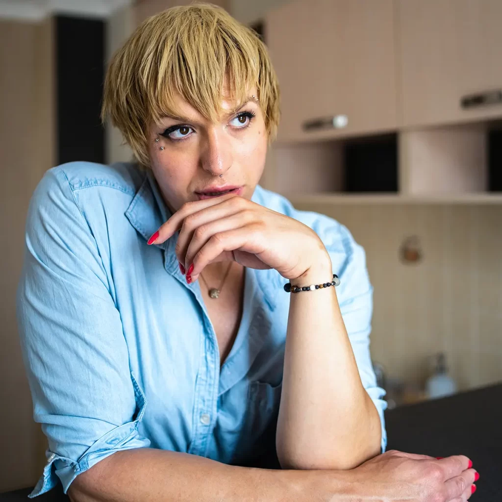 Young transgender person with blonde hair and blue shirt, sitting at table with hand on chin.